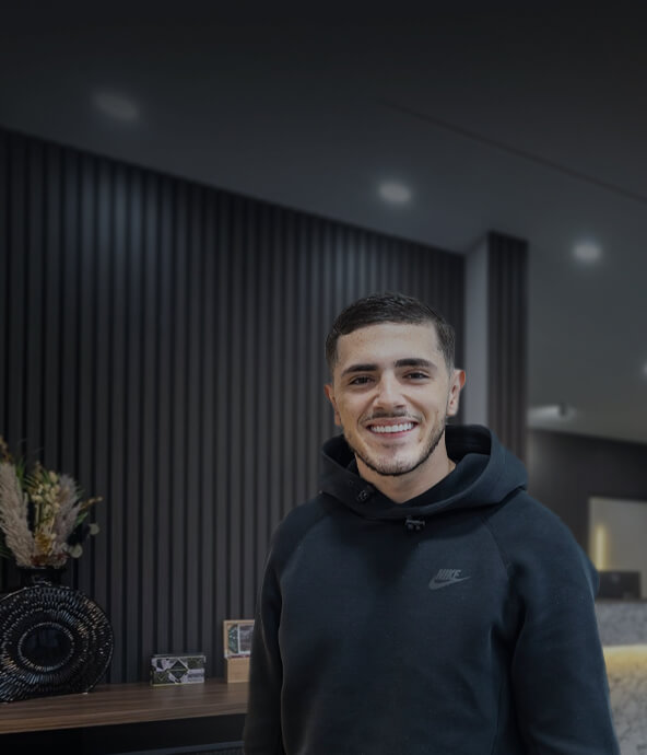 A young man wearing a black hoodie with a Nike logo stands smiling in front of a modern, textured wall.