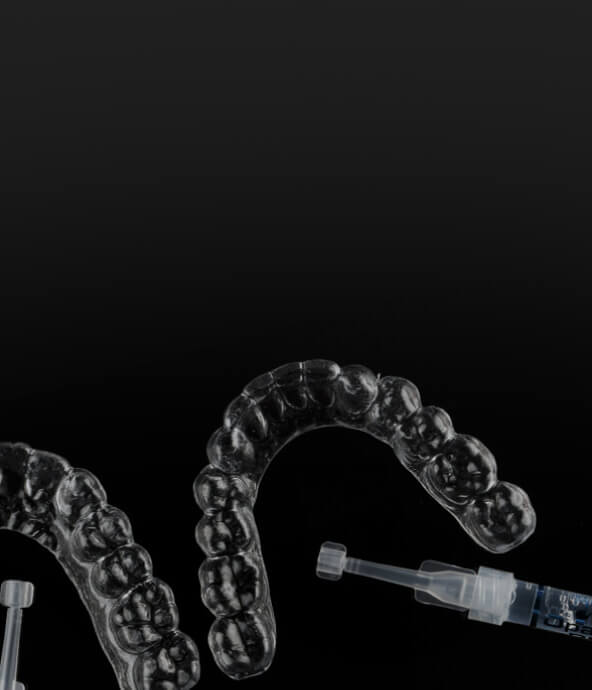A close-up image of clear dental aligners placed on a black background, with syringes beside them.