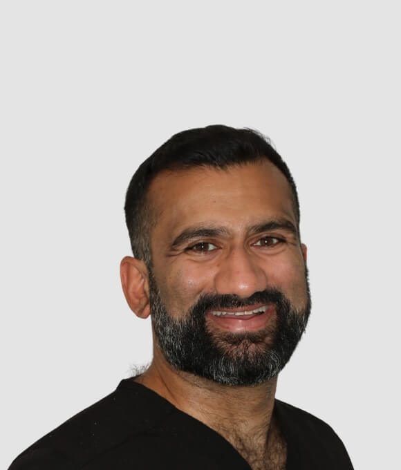 A smiling male dentist with a beard is wearing a black medical scrub, facing the camera with a neutral background.