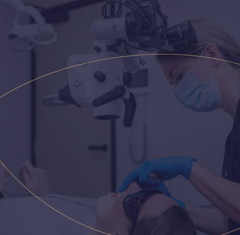 Image of a dental professional using a microscope while performing treatment on a patient, with the patient wearing protective glasses.