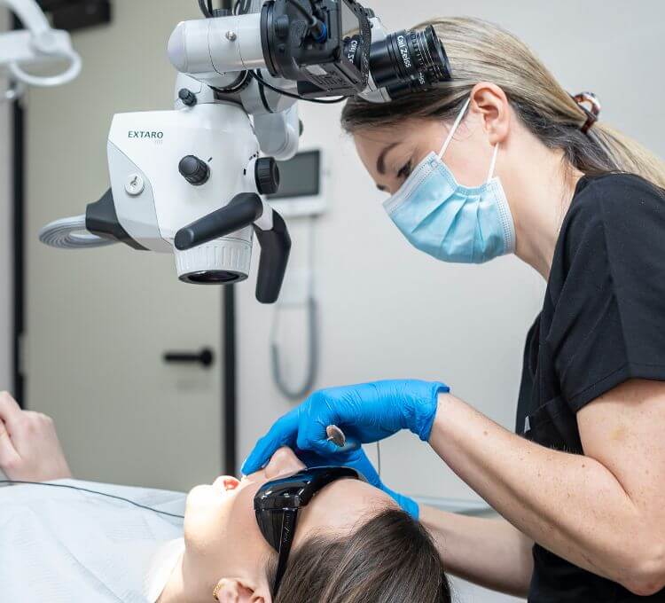 A dentist wearing a mask and gloves uses a microscope while performing dental treatment on a patient who is wearing protective glasses.