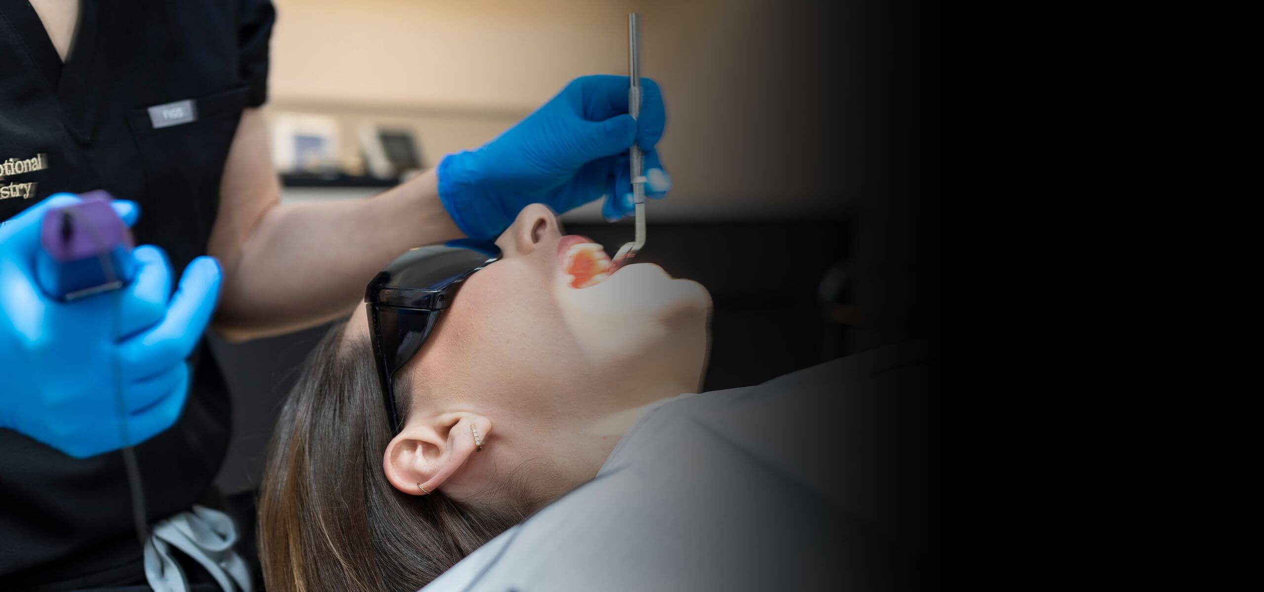 Dentist performing a treatment on a patient, using dental tools while the patient wears protective glasses.